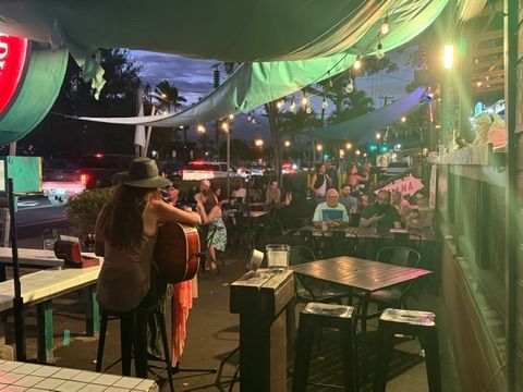 Kris Bradley performing live with acoustic guitar at an outdoor Maui restaurant patio, string lights overhead.